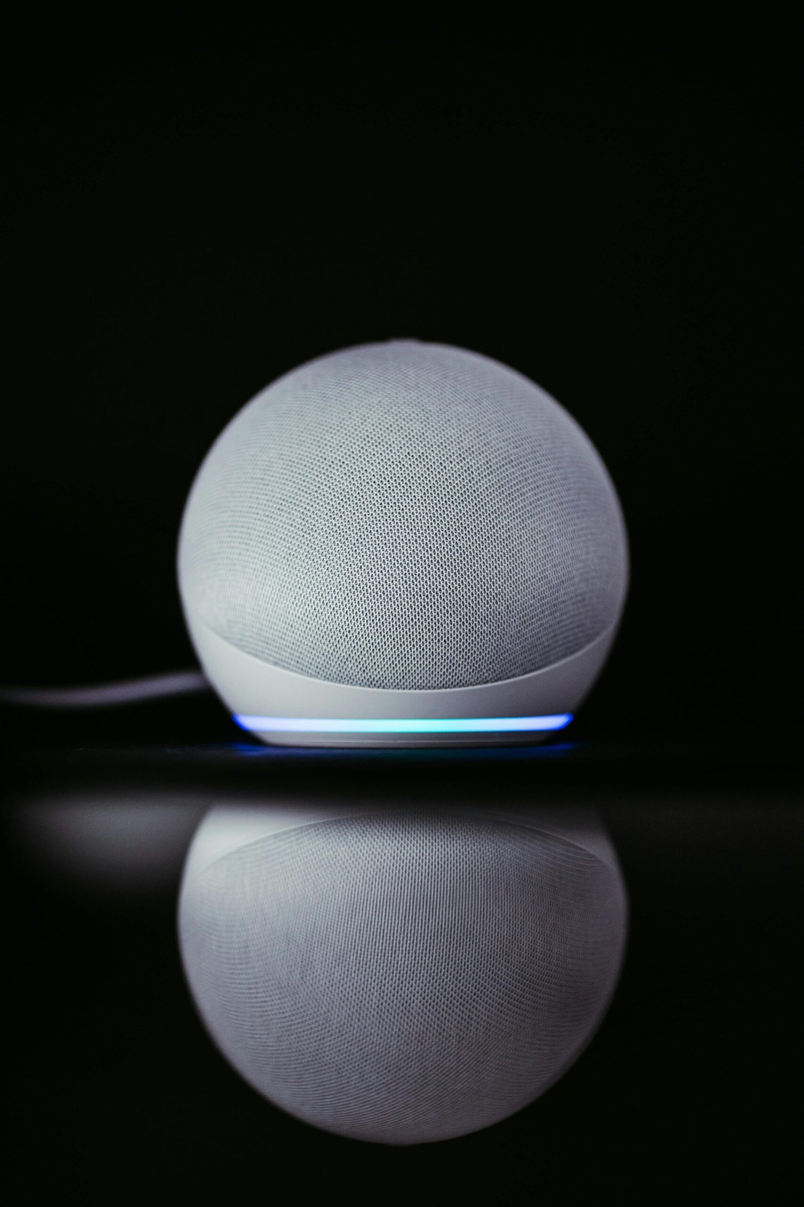 A modern smart speaker Echo Dot with a sleek design and glowing base, reflected on a dark surface.